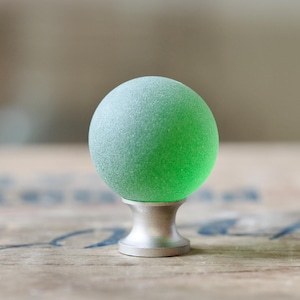 Peut inclure: Un bouton de porte en verre vert avec une base argentée. Le bouton est rond et a une finition givrée.