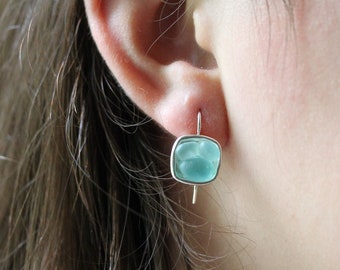 May include: A silver earring featuring a square, pale blue, sea glass-style stone. The earring is suspended from the ear. The stone is set in a silver frame and has a slightly textured appearance.