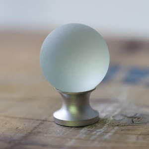 May include: A spherical cabinet knob featuring a frosted, translucent glass ball top attached to a smooth, brushed silver-toned metal base. The hardware has a clean, rounded design with a matte finish on the metal component and a soft, diffused appearance on the glass sphere.