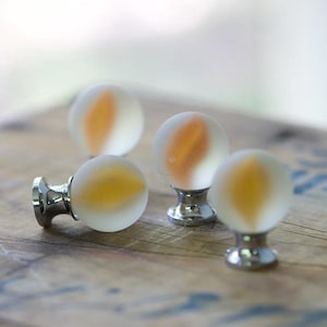 Può includere: Quattro pomelli sferici per mobili con finitura in vetro bianco satinato e un motivo centrale a forma di fiamma color ambra. Ogni pomello è fissato su una base in metallo color argento lucido, progettata per cassetti o ante di armadi.