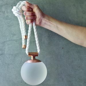 Puede incluir: Una lámpara colgante de cuerda blanca con una esfera de vidrio esmerilado y detalles en cobre.