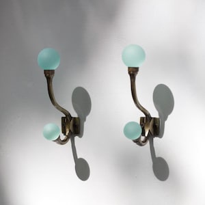 Peut inclure: Deux patères murales composées d'une structure en métal doré courbé à deux bras. Chaque bras est surmonté d'un bouton sphérique en verre dépoli vert menthe clair. La base métallique est carrée et présente des vis de fixation visibles.