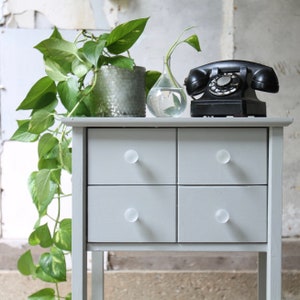 May include: A gray nightstand with four drawers and clear glass knobs. The nightstand has a vintage rotary phone on top, a potted plant, and a clear glass vase with a plant.