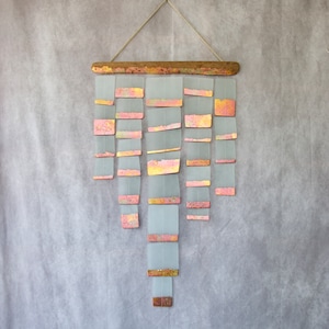 May include: A wall hanging made of driftwood and glass squares. The glass squares are translucent and have a pink and gold metallic finish. The squares are arranged in a cascading pattern.