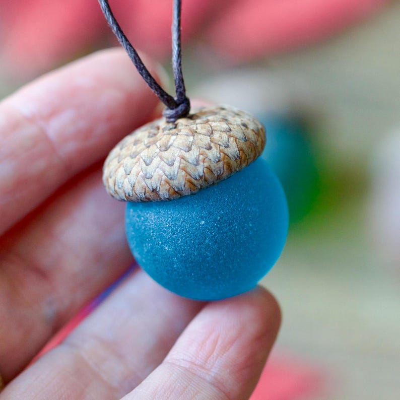 May include: A unique acorn pendant featuring a vibrant blue glass orb and a natural acorn cap. The pendant is suspended from a dark brown cord. The blue orb is approximately 1 inch in diameter, creating a striking contrast with the natural acorn cap.