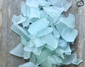 May include: A pile of sea glass pieces in various shapes and sizes. The glass has a frosted, translucent appearance with shades of light blue and aqua. The pieces are scattered on a weathered, gray wooden surface.