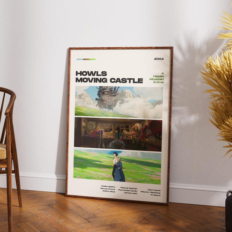 May include: A framed poster for the animated film "Howl's Moving Castle" by Hayao Miyazaki. The poster features three scenes from the movie, with the title and year 2004 at the top. The poster is in a wooden frame and leans against a white wall.