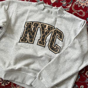 May include: A light gray long-sleeve sweatshirt featuring a mock neck collar and ribbed cuffs. The center of the chest displays the letters NYC in a bold, block-style font with a brown and black leopard print pattern and a thin black outline.