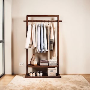 Peut inclure: Un portant à vêtements en bois brun foncé avec divers vêtements suspendus. Le portant a une étagère avec un sac à main, des vêtements pliés et une petite boîte. En dessous, des paires de chaussures. Le sol est recouvert d'un tapis moelleux.