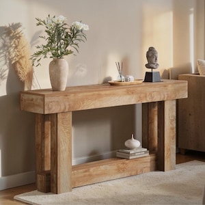 Op de afbeelding: Een lichtbruine houten console met een rechthoekige bovenkant en twee verticale steunen. De tafel is versierd met een vaas met witte bloemen, een kleine Boeddha-beeld en andere decoratieve items. De tafel staat op een lichtgekleurd tapijt.