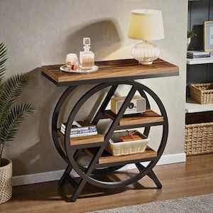 May include: A contemporary side table with a dark wood top and shelves, supported by a black metal frame with a circular design. The table holds a lamp, decanter, glasses, books, a radio, and a basket. The measurements are in centimeters.