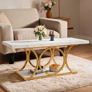 May include: A rectangular coffee table with a white marble-effect top and gold-coloured metal base. The base has a circular design. A lower shelf and a basket of oranges are visible. The table is set on a beige rug.