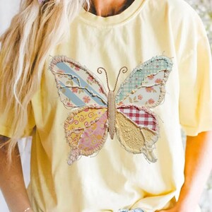 May include: A pale yellow t-shirt with a large butterfly appliqué. The butterfly is made from assorted patterned fabrics in blue, pink, red, and yellow. The shirt has a round neck and short sleeves. The butterfly design is approximately 20 cm wide.