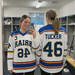 May include: A white hockey jersey with blue and yellow stripes on the sleeves and hem. The front features the text BRIAR and the number 46 in blue with yellow outlines. The back displays the name TUCKER and the number 46 in blue and yellow.