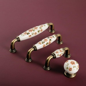 Puede incluir: Juego de herrajes para armarios con tres tiradores y un pomo. Los tiradores tienen un acabado cerámico blanco con un estampado floral en naranja y verde. Los herrajes tienen un acabado bronce, sobre fondo burdeos.
