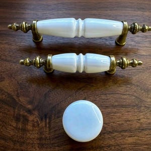 Puede incluir: Dos tiradores de armario de cerámica blanca con herrajes color latón y un pomo redondo a juego. Los tiradores tienen un diseño clásico con un detalle central acanalado. Los herrajes tienen un acabado antiguo, sobre una superficie de madera.