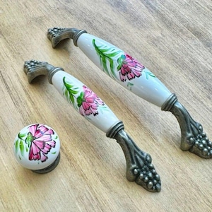 May include: Two white ceramic cabinet handles and a knob with a floral design in pink and green. The handles have a silver-colored metal base. The knob is round and has a matching floral design.