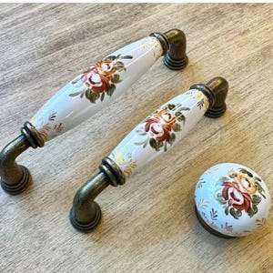May include: Set of three decorative cabinet hardware pieces. Two elongated handles and a round knob feature a white ceramic finish with hand-painted floral designs in shades of red, orange, and green. The hardware has a bronze-colored metal base.