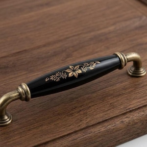 Op de afbeelding: Zwarte en gouden kastgreep met een bloemmotief. De handgreep is zwart met gouden bloemaccenten en goudkleurige metalen hardware. De greep is gemonteerd op een houten oppervlak.