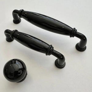 May include: A set of black cabinet hardware, including two pull handles and a round knob. The handles have a curved design with decorative detailing. The hardware is glossy and reflects light, suggesting a polished finish. The items are arranged on a white surface.
