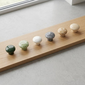 Könnte beinhalten: Sechs dekorative Schrankknöpfe aus Stein in verschiedenen Farben und Texturen. Sie sind auf einer hellen Holzplatte montiert. Die Knöpfe sind in Grün-, Weiß-, Grau- und Beigetönen gehalten. Jeder Knopf hat eine abgerundete Form.