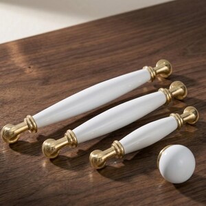 May include: A collection of white and gold cabinet hardware, including three pull handles and a round knob. The handles have a white ceramic center and gold-toned metal ends. The hardware is arranged on a wooden surface.