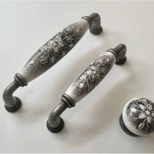 May include: Set of decorative cabinet hardware featuring two pull handles and a knob. The handles and knob have a white and gray floral design on a ceramic surface, with dark gray metal bases and accents. The hardware is designed for cabinets and drawers.