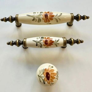 May include: A set of cream-coloured cabinet handles and a knob. The handles and knob feature a hand-painted floral design in orange, brown, and green. The metal accents are antique brass-toned. The handles are approximately 15 cm long.
