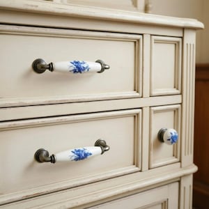 May include: Close-up of a cream-colored dresser with decorative drawer pulls. The pulls are white ceramic with blue floral designs and antique bronze metal hardware. The dresser has a distressed finish, giving it a vintage look.
