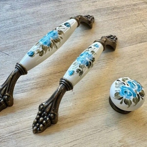 May include: Two cabinet pulls and a knob. The pulls have a white ceramic center with blue floral designs and bronze-colored metal hardware. The knob has a matching design. The hardware has a decorative grape cluster design.