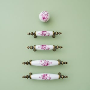 May include: A collection of white ceramic cabinet hardware with a floral design in shades of pink and purple. The hardware includes four pull handles and one round knob, all with brass-colored metal accents. The items are arranged vertically against a light green background.