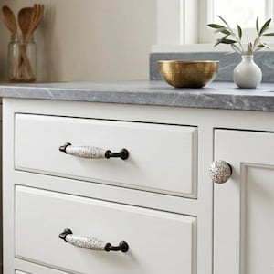 Può includere: Armadietti da cucina bianchi con ferramenta decorativa. I cassetti hanno lunghe maniglie con finitura craquelé con dettagli in metallo nero. Un pomello abbinato con finitura craquelé è sulla porta dell'armadio. Il piano di lavoro è in pietra grigia.