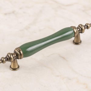 May include: A cabinet pull with a dark green ceramic handle and antique brass-colored metal accents. The handle is cylindrical, and the metal base has a decorative, ornate design. The hardware is designed for furniture.