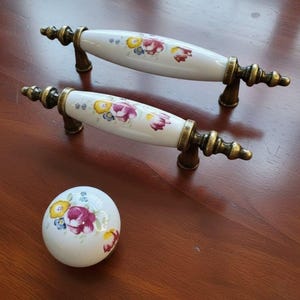 May include: Two white ceramic cabinet handles and a matching knob. The handles feature a floral design in yellow, purple, and red, with bronze-coloured metal hardware. The knob has a similar floral pattern.