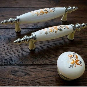 May include: A set of cabinet hardware featuring two white and gold floral-patterned handles and a matching knob. The handles have silver-colored metal bases and decorative ends. The knob is round with a similar floral design and silver base. The hardware is displayed on a dark wood surface.