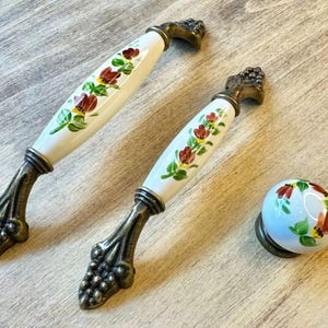 May include: Two white ceramic cabinet handles and a knob with a floral design. The handles have a bronze-colored base with a grape cluster design. The floral pattern includes red and yellow flowers with green leaves.