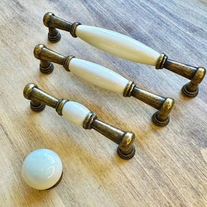 May include: Three antique-style cabinet pulls and a knob. The pulls have a bronze finish with white ceramic center sections. The knob is white ceramic with a bronze base. Ideal for kitchen or bathroom cabinets.