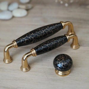May include: Set of two black and gold cabinet pulls and a matching knob. The pulls have a gold base and a black, patterned handle. The knob is round with a similar black and gold pattern. Hardware for furniture.