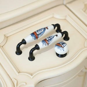May include: Set of cabinet hardware featuring two long handles and a knob. The handles and knob have a white ceramic finish with a nautical design in blue and orange. The hardware has black metal bases.
