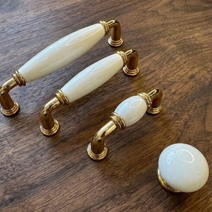 Op de afbeelding: Een set meubelbeslag met witte keramische handgrepen en goudkleurige metalen details. De set bevat drie trekhandgrepen en een knop, allemaal met een klassiek ontwerp. Gepresenteerd op een donkerhouten oppervlak, geschikt voor kasten.
