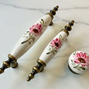 May include: Set of three decorative cabinet hardware pieces. Includes two white ceramic handles and one knob, all featuring a pink flower design with green leaves. The hardware has antique brass-colored metal accents.