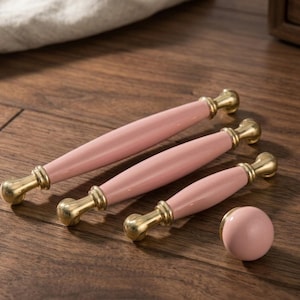 Op de afbeelding: Een set van vier meubelbeslagstukken: drie roze keramische handgrepen en een roze knop. De handgrepen hebben een goudkleurige basis en uiteinden. De knop is rond. Alle stukken liggen op een houten oppervlak.