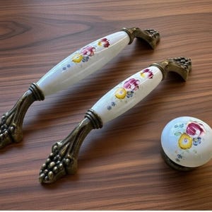 Puede incluir: Dos tiradores de armario de cerámica blanca y un pomo a juego con diseños florales en rojo, amarillo y morado. Los tiradores tienen manijas de metal ornamentadas de color bronce. El pomo es redondo con un patrón floral similar.
