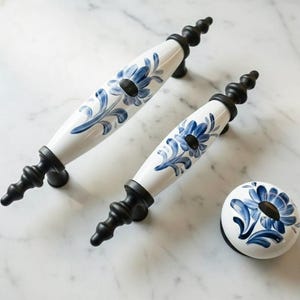 Puede incluir: Tiradores y pomo de armario de cerámica floral blanca y azul con herrajes negros. Los tiradores tienen un diseño curvo con un estampado floral azul. El pomo es redondo con un diseño floral a juego.