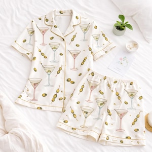 May include: White pajama set with a martini and olive print. The short-sleeved top and shorts feature a repeating pattern of cocktail glasses and olive skewers. The set has gold trim.