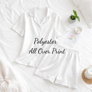 Puede incluir: Conjunto de pijama blanco de poli&eacute;ster con una camisa de manga corta con botones y pantalones cortos a juego. El conjunto presenta un cuello cl&aacute;sico y lleva la etiqueta "Polyester All Over Print". El conjunto se muestra en una cama blanca.