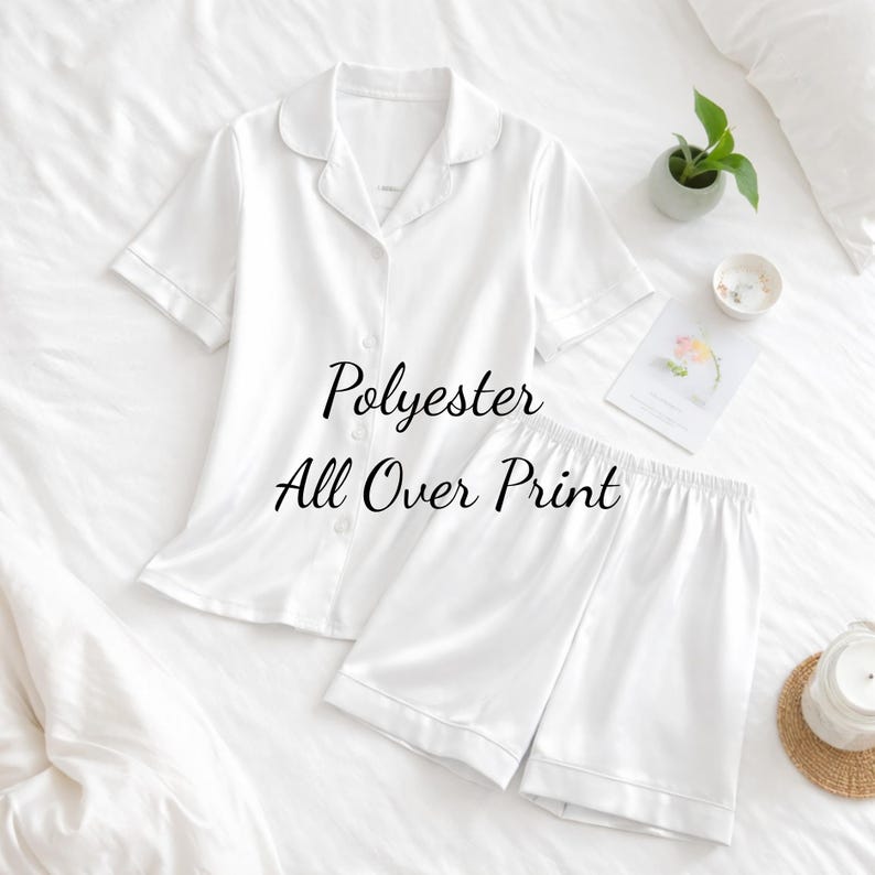 Puede incluir: Conjunto de pijama blanco de poli&eacute;ster con una camisa de manga corta con botones y pantalones cortos a juego. El conjunto presenta un cuello cl&aacute;sico y lleva la etiqueta "Polyester All Over Print". El conjunto se muestra en una cama blanca.