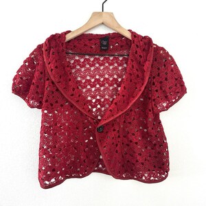 May include: A short-sleeved red crochet bolero jacket featuring an open-front design with a single black button closure, a wide collar, and a delicate, repeating floral-inspired lace pattern throughout the entire garment.