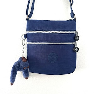 Könnte beinhalten: Eine dunkelblaue Umhängetasche mit zwei horizontalen Reißverschlusstaschen auf der Vorderseite, silberfarbenen Metallreißverschlüssen und einem runden Logo-Aufnäher. Ein flauschiger blauer Affen-Schlüsselanhänger hängt an einem Metallring.