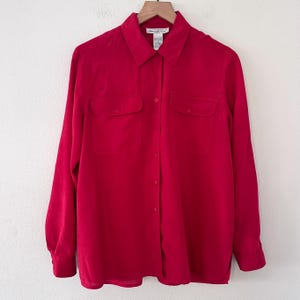 May include: A vibrant red button-down shirt with long sleeves and two chest pockets. The shirt is hanging on a wooden hanger against a plain white background. The shirt has a classic collar and buttoned cuffs.
